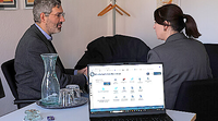 Laptop mit Förderportal und Gespräch zwischen zwei Personen