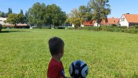 Kind hat einen Ball in der Hand und schaut auf einen Sportplatz