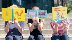 Drei Menschen halten sich ein Buch vor das Gesicht und lesen