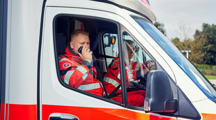 Ein Mitarbeiter aus dem Rettungsdienst fährt mit seiner Kollegin im Rettungswagen und spricht in ein Funkgerät.